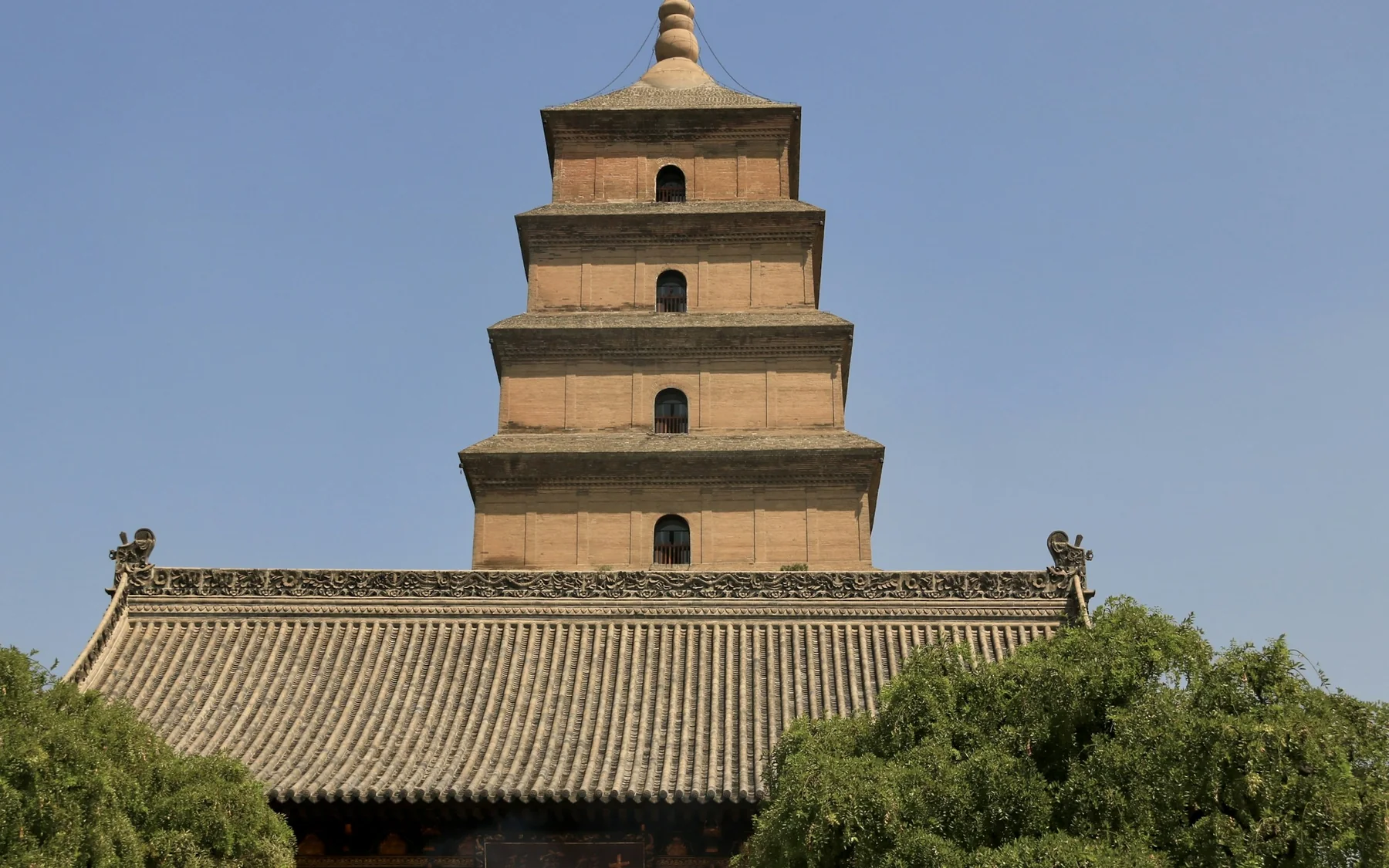 The brick Giant Wild Goose Pagoda rising above the tiled roof and trees of Da Ci'en Temple in Xi'an under a clear blue sky.