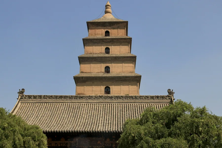 The brick Giant Wild Goose Pagoda rising above the tiled roof and trees of Da Ci'en Temple in Xi'an under a clear blue sky.