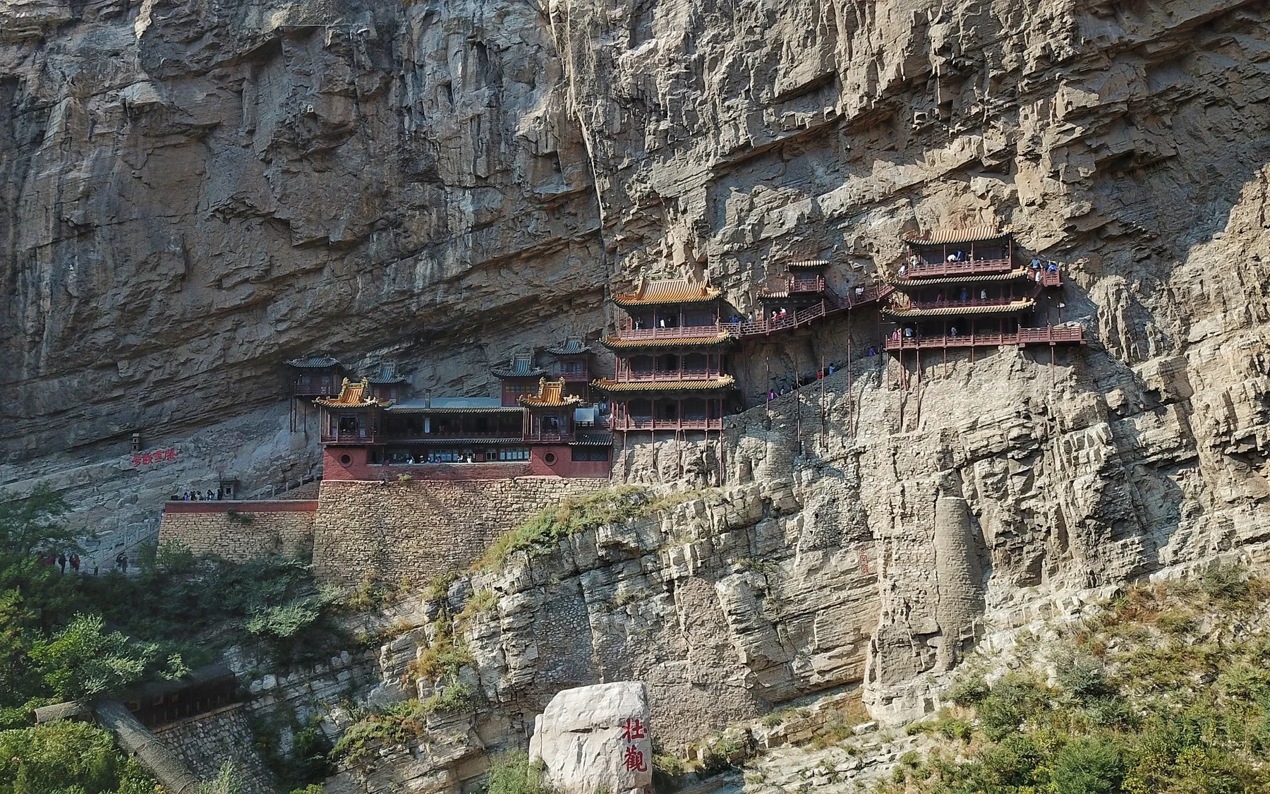 A wide aerial view of the real Hanging Temple at Hunyuan, its timber halls and galleries clinging to the pale cliff above the gorge.