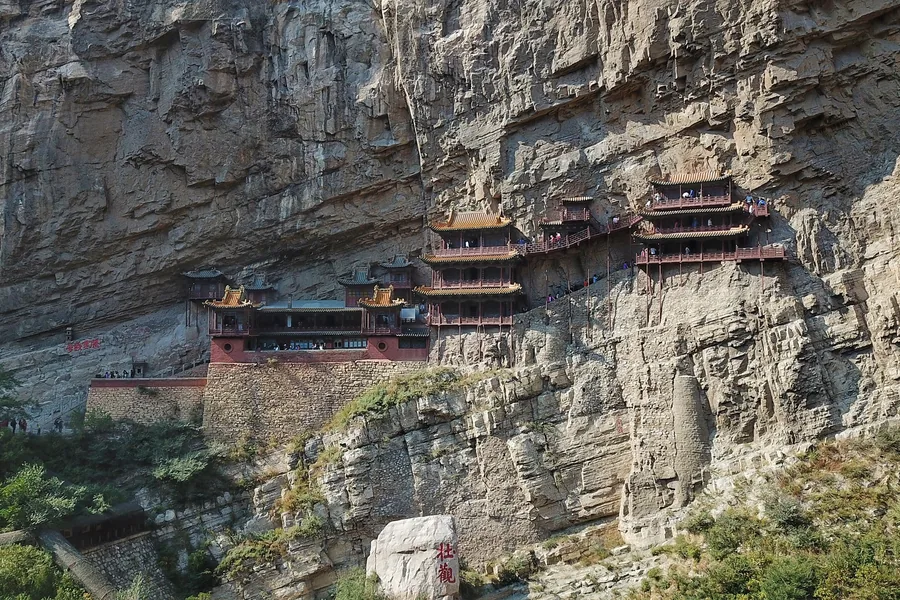 A wide aerial view of the real Hanging Temple at Hunyuan, its timber halls and galleries clinging to the pale cliff above the gorge.