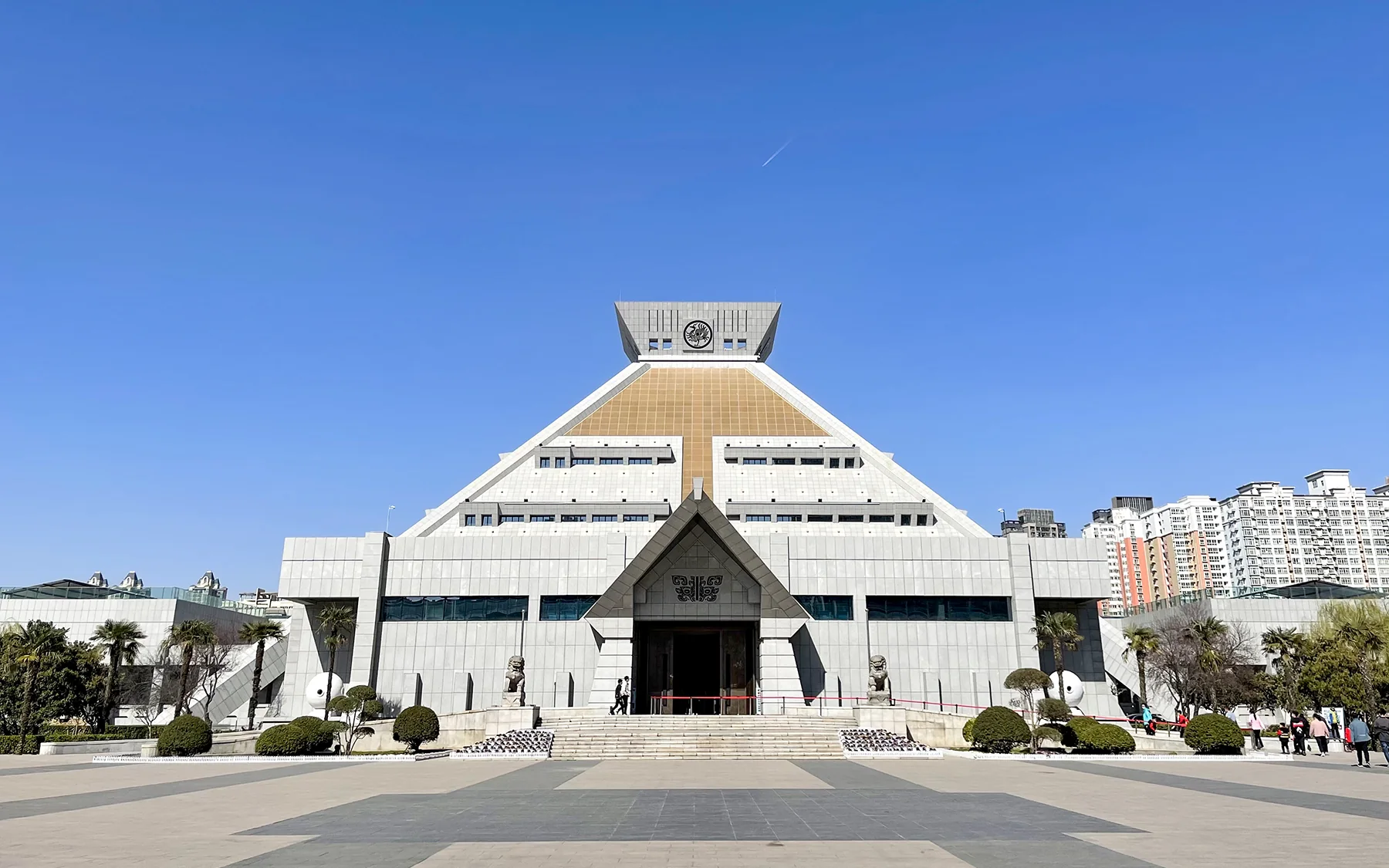 A clear frontal view of Henan Museum's complete stepped-pyramid main hall, centred across its forecourt beneath a deep blue sky.