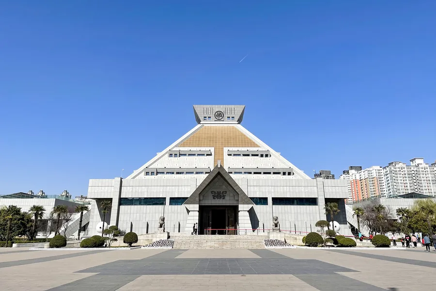 A clear frontal view of Henan Museum's complete stepped-pyramid main hall, centred across its forecourt beneath a deep blue sky.