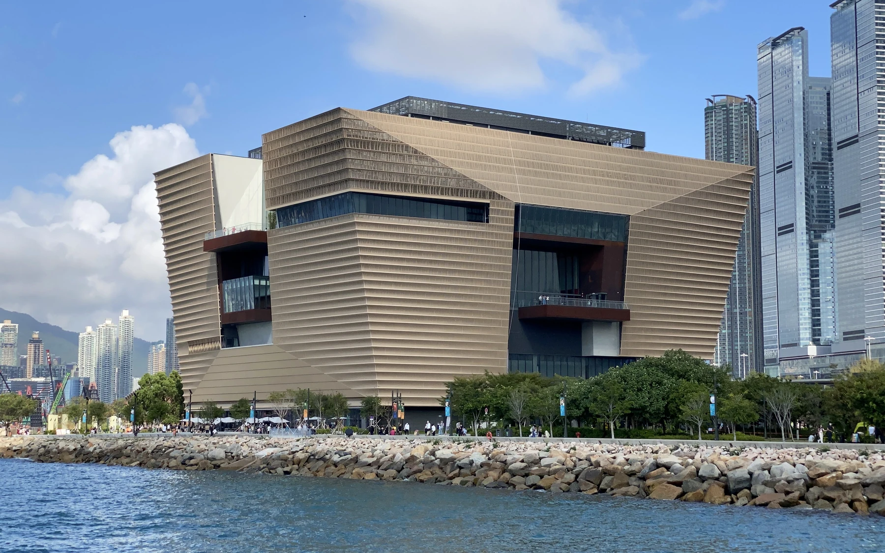 The bronze-coloured Hong Kong Palace Museum beside Victoria Harbour under a clear blue sky.