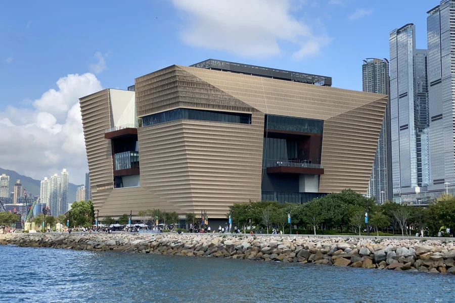The bronze-coloured Hong Kong Palace Museum beside Victoria Harbour under a clear blue sky.