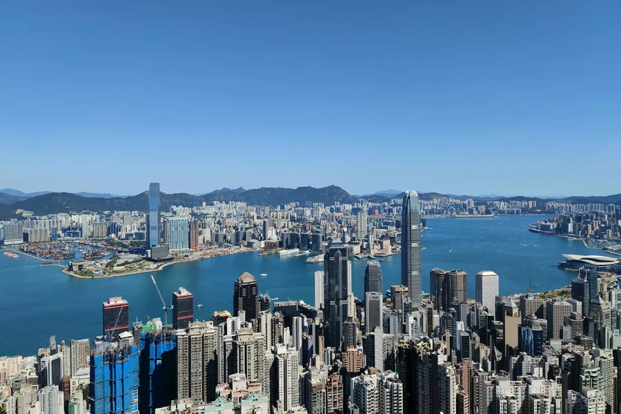 A clear daytime panorama from Victoria Peak across central Hong Kong and Victoria Harbour toward the Kowloon skyline, with the harbour forming the visual centre.