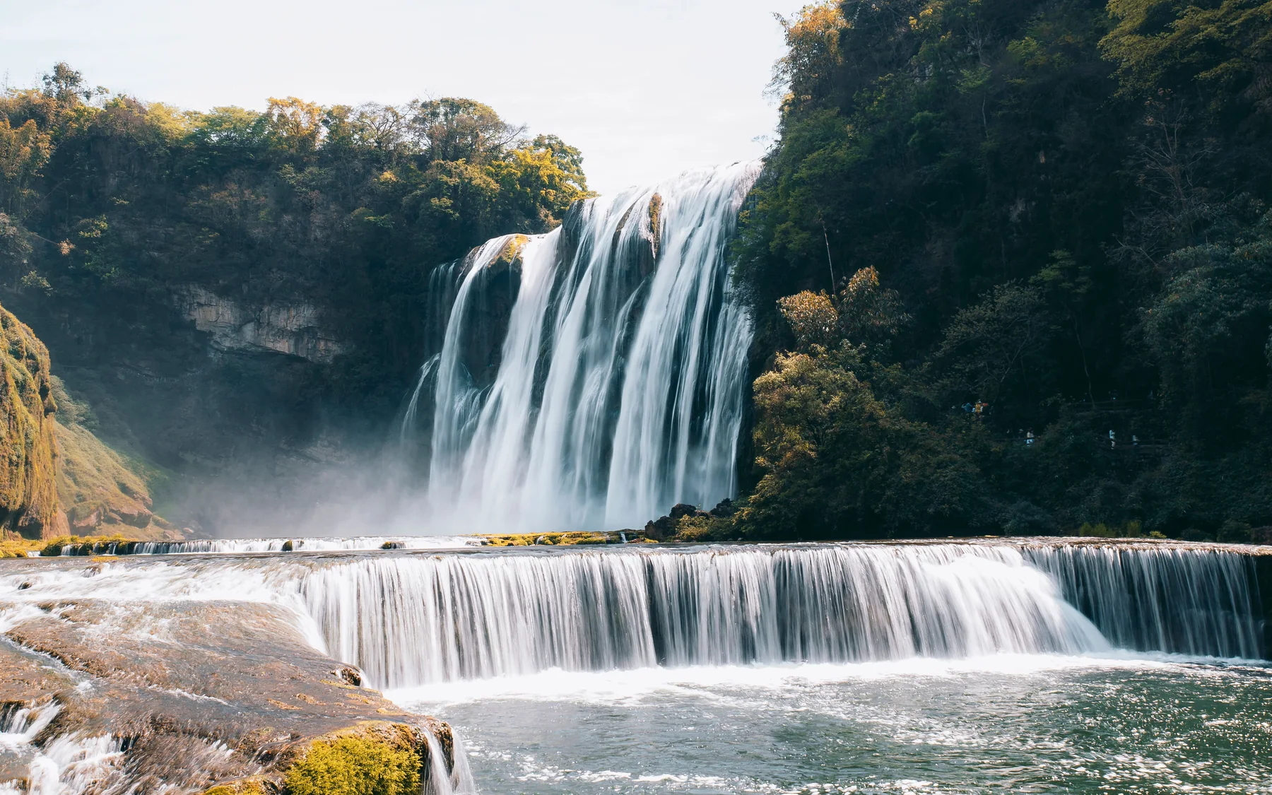 The broad white curtain of Huangguoshu Waterfall cascading into a misty river amid forested cliffs in Guizhou.