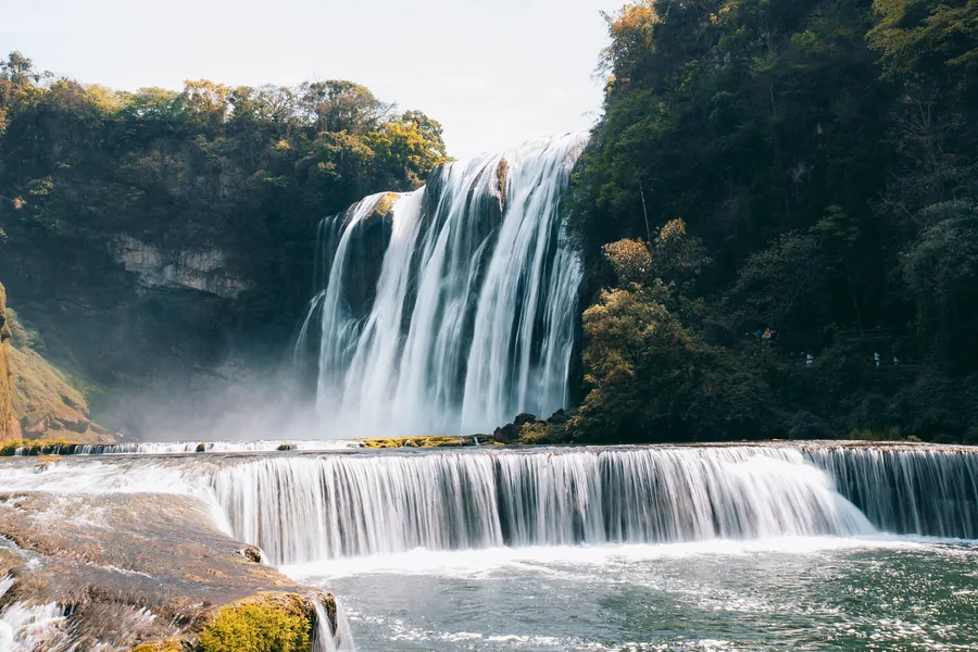 The broad white curtain of Huangguoshu Waterfall cascading into a misty river amid forested cliffs in Guizhou.
