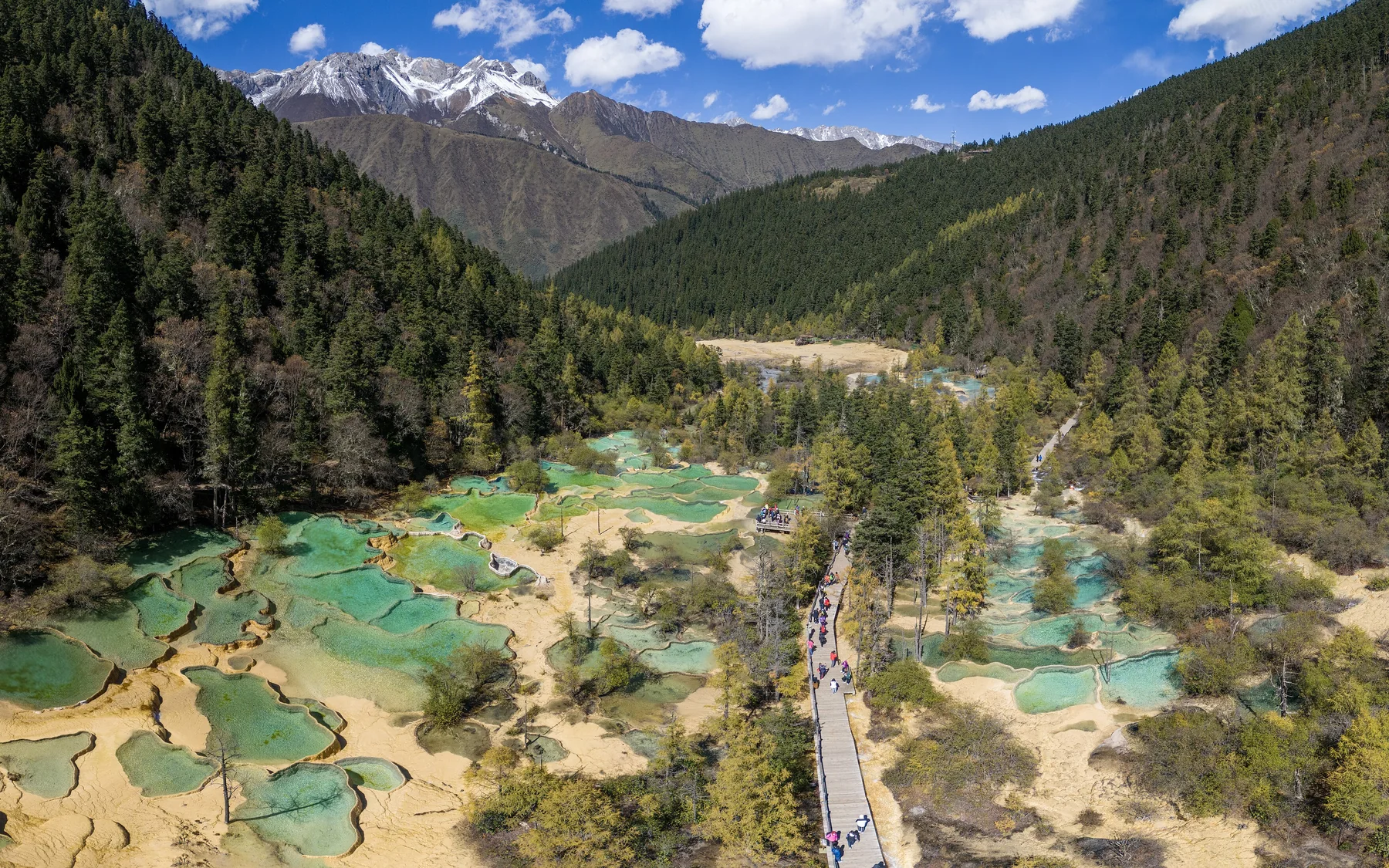 An aerial panorama of Huanglong's turquoise travertine pools descending through a forested valley toward snow-capped mountains.