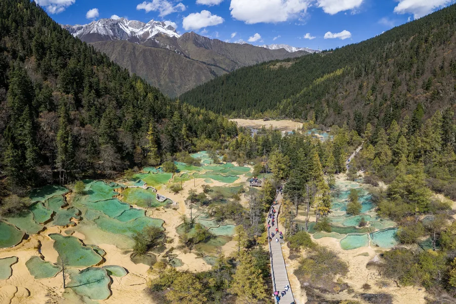 An aerial panorama of Huanglong's turquoise travertine pools descending through a forested valley toward snow-capped mountains.