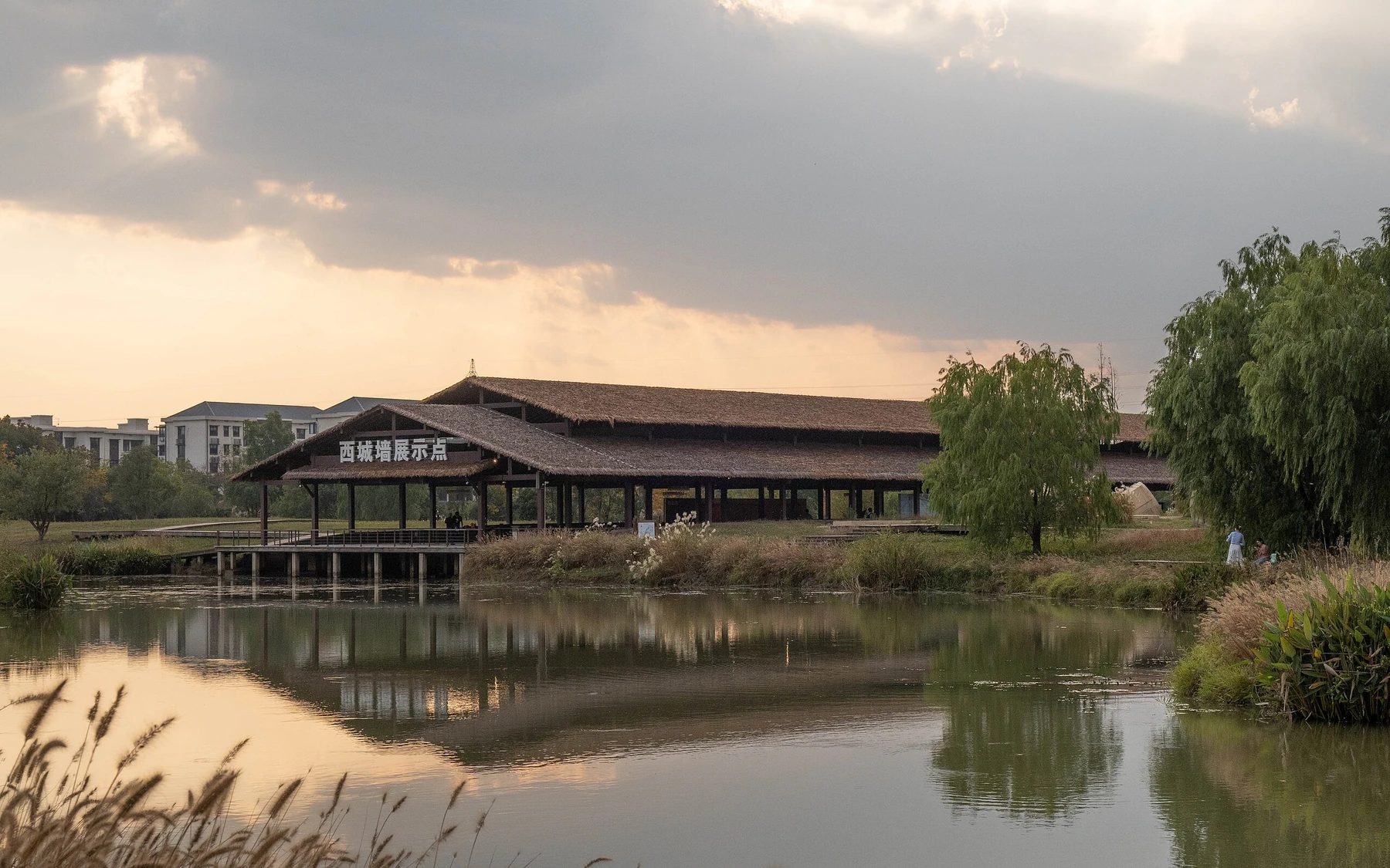 The West City Wall display area at Liangzhu Archaeological Park reflected in water beneath a dramatic evening sky.