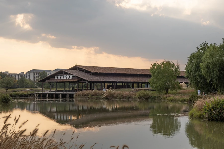 The West City Wall display area at Liangzhu Archaeological Park reflected in water beneath a dramatic evening sky.