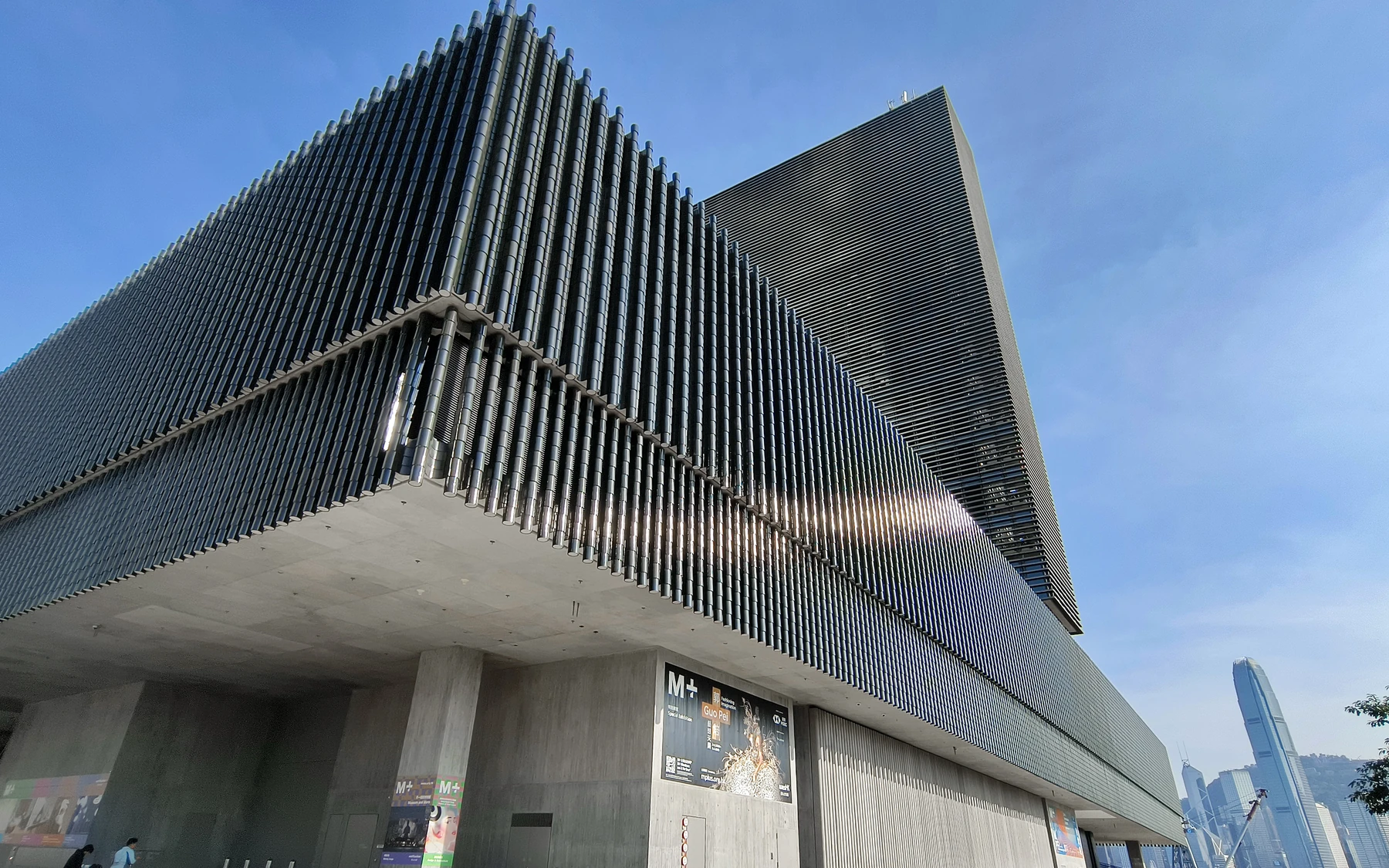 The dark ceramic-tile facade and projecting gallery volume of M+ in West Kowloon beneath a clear blue sky.