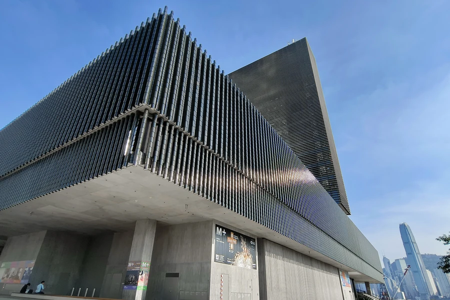 The dark ceramic-tile facade and projecting gallery volume of M+ in West Kowloon beneath a clear blue sky.