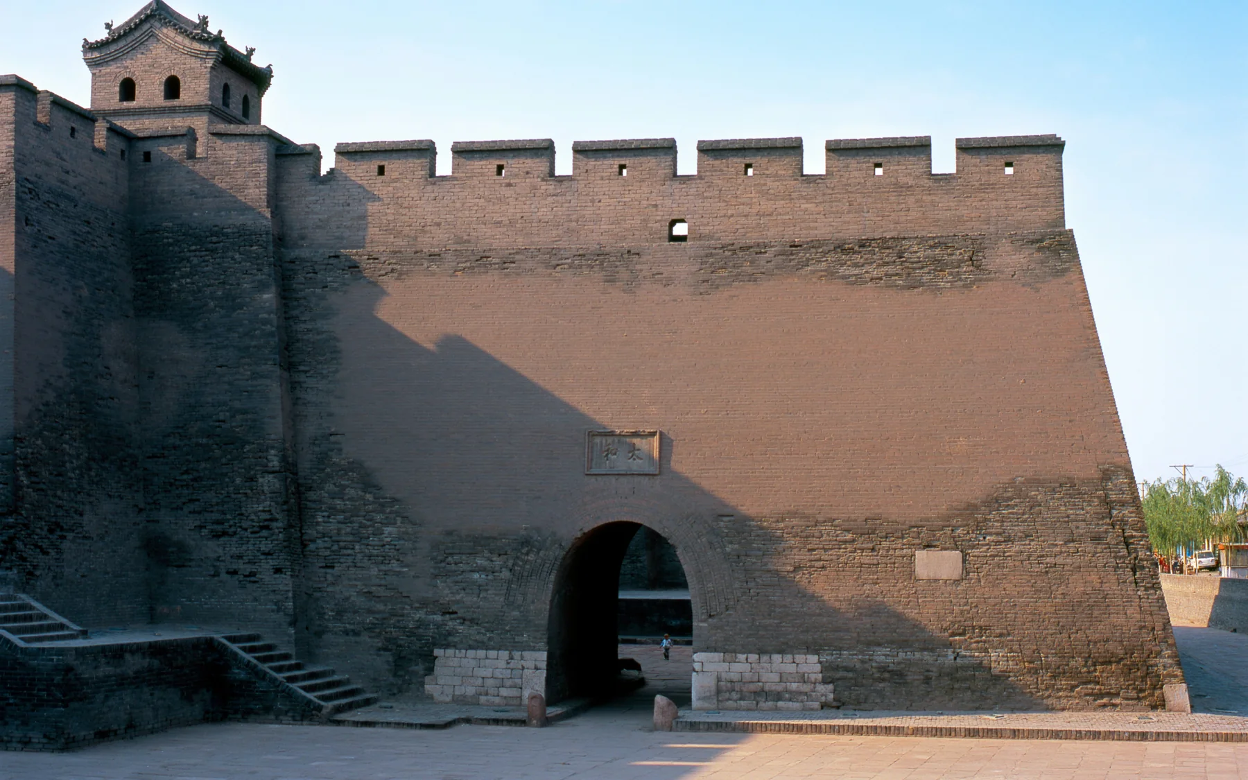 A crenellated gate tower and massive grey-brick defensive wall in Pingyao Ancient City under a clear blue sky.