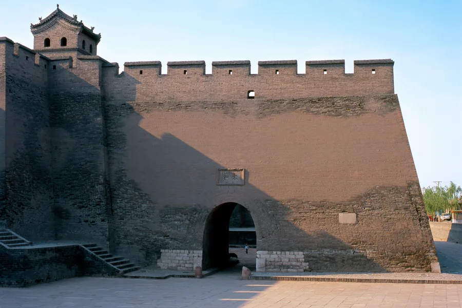 A crenellated gate tower and massive grey-brick defensive wall in Pingyao Ancient City under a clear blue sky.