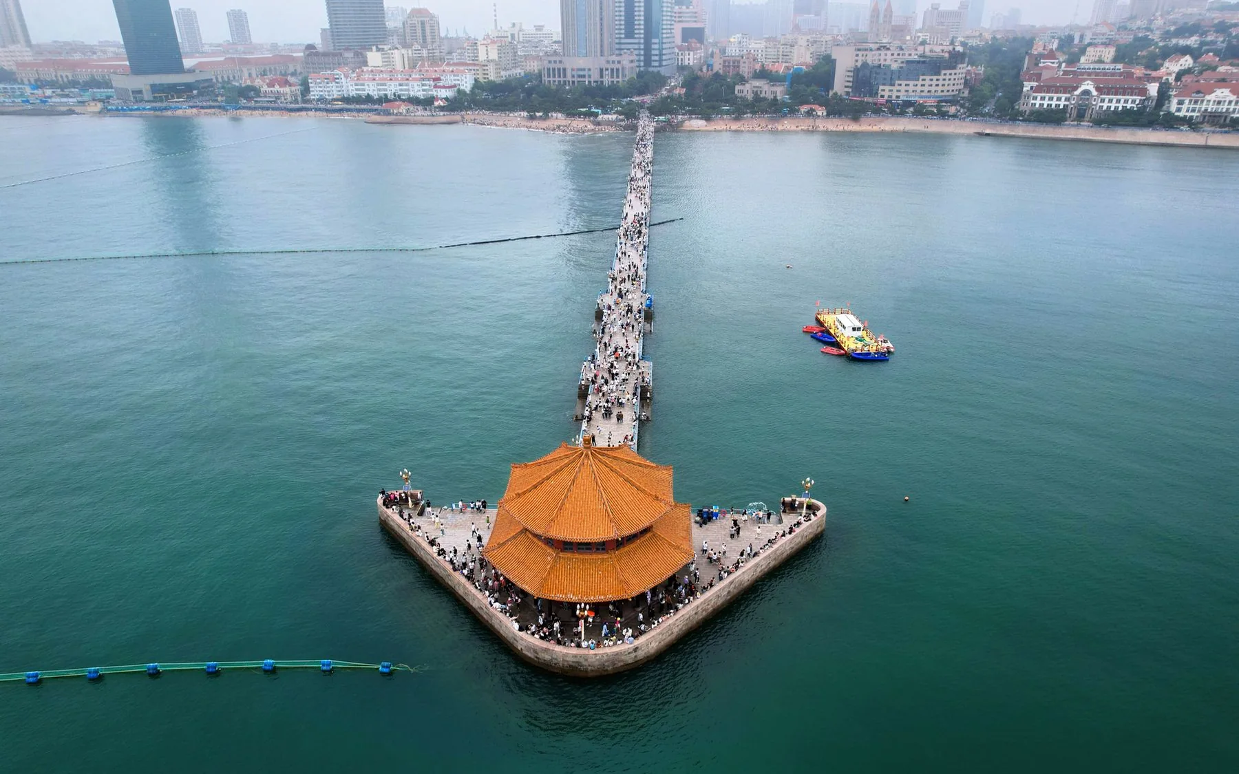 An aerial view along Qingdao's Zhanqiao Pier, with the orange-roofed Huilan Pavilion surrounded by the sea and the historic city rising behind the shoreline.