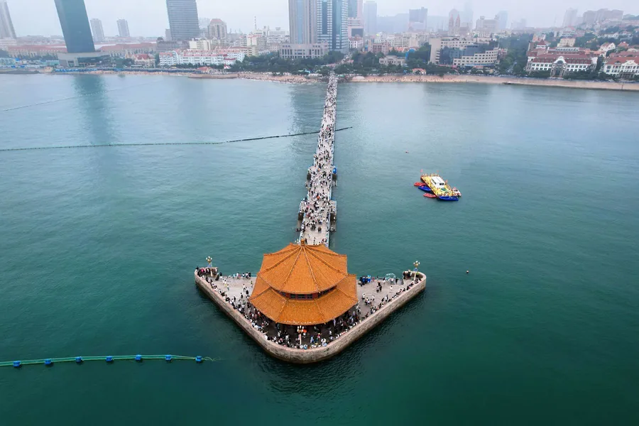 An aerial view along Qingdao's Zhanqiao Pier, with the orange-roofed Huilan Pavilion surrounded by the sea and the historic city rising behind the shoreline.