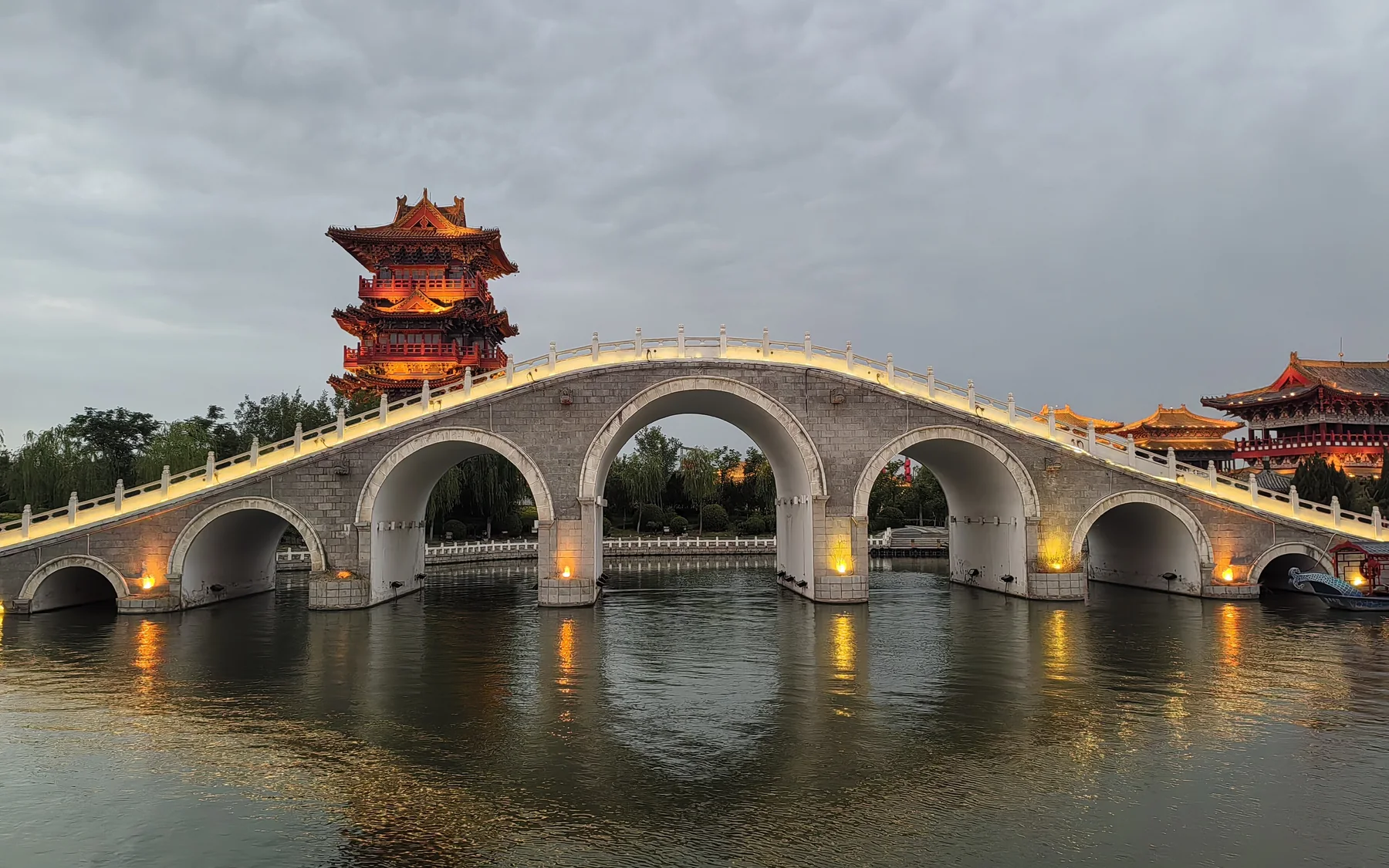 Jiulong Bridge at Qingming Riverside Landscape Garden, illuminated across the water with a multi-storey Song-style tower and waterfront buildings behind it.