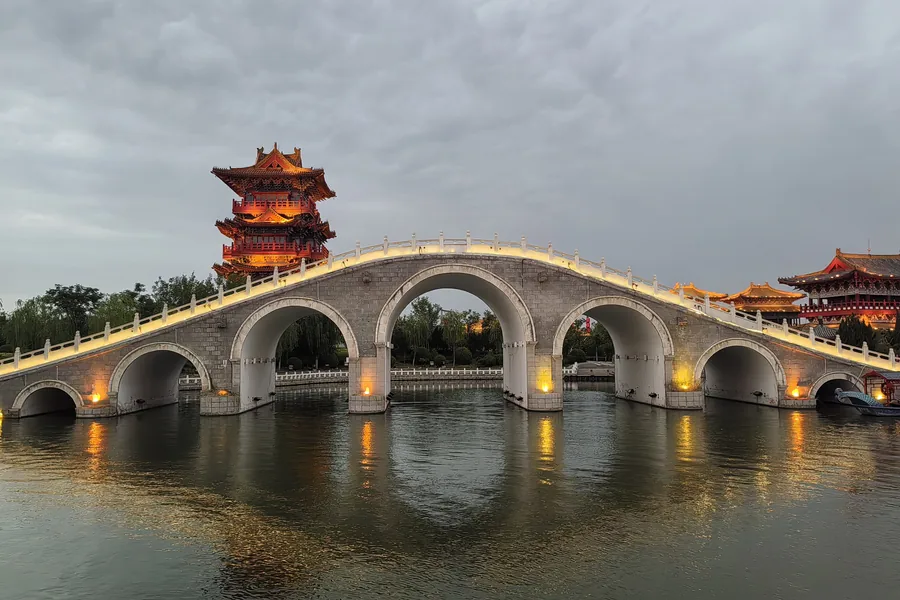 Jiulong Bridge at Qingming Riverside Landscape Garden, illuminated across the water with a multi-storey Song-style tower and waterfront buildings behind it.