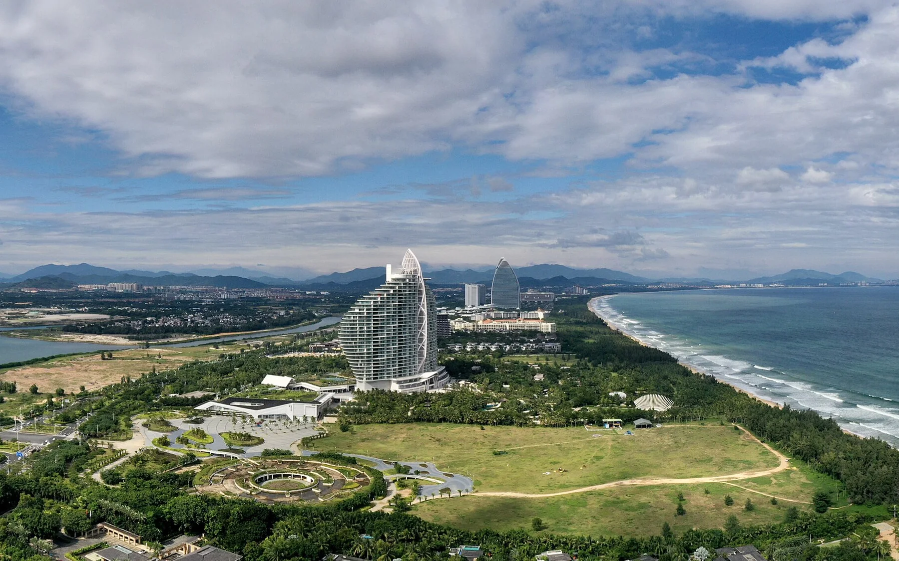 A high, photographer-composed panorama of Haitang Bay in Sanya, with the full tropical shoreline, blue-green sea, beachfront resorts and mountain backdrop visible in clear daylight.