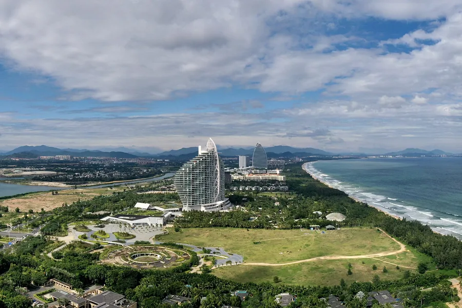 A high, photographer-composed panorama of Haitang Bay in Sanya, with the full tropical shoreline, blue-green sea, beachfront resorts and mountain backdrop visible in clear daylight.