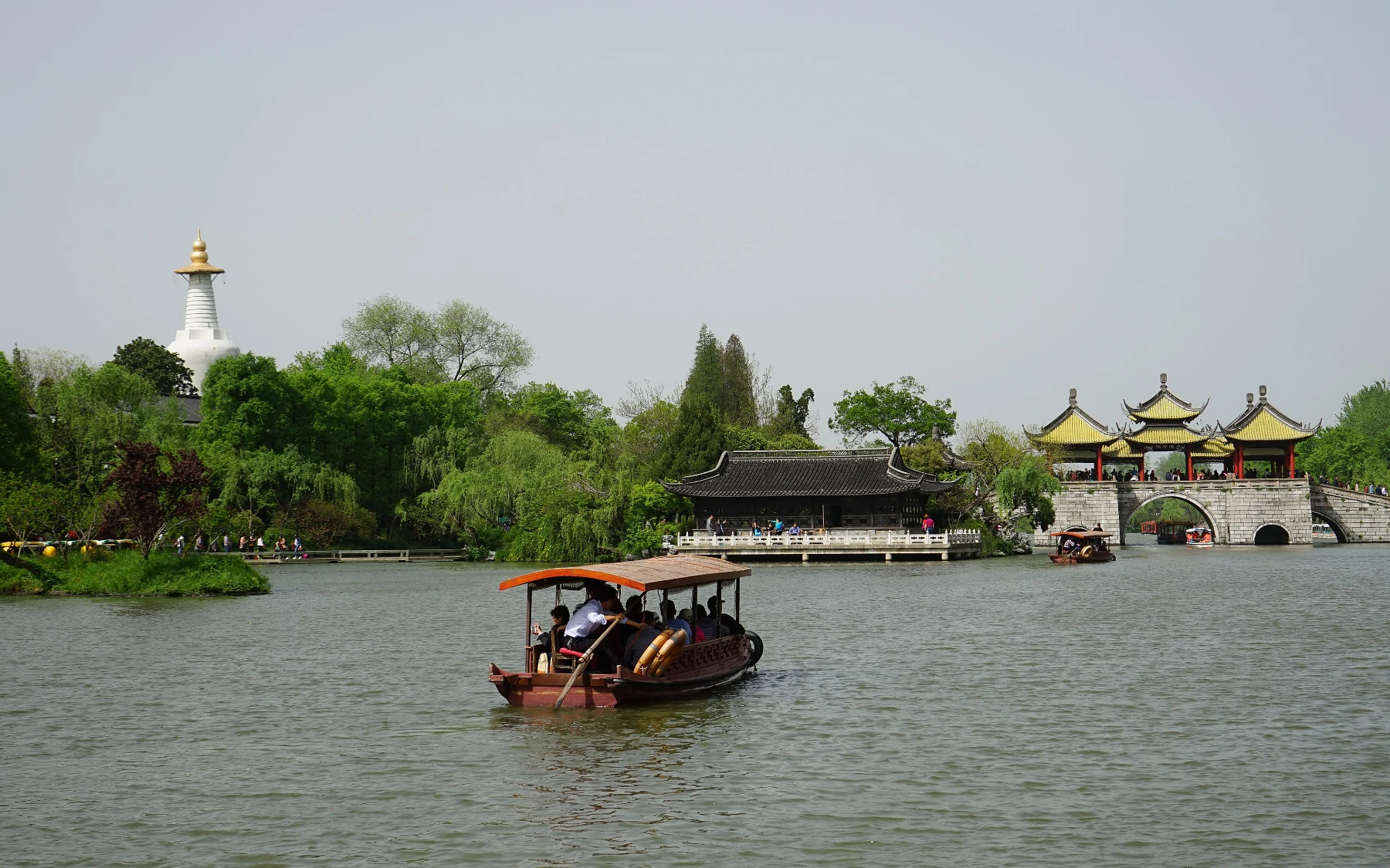 A wooden tour boat crossing Slender West Lake between the White Pagoda and Five-Pavilion Bridge