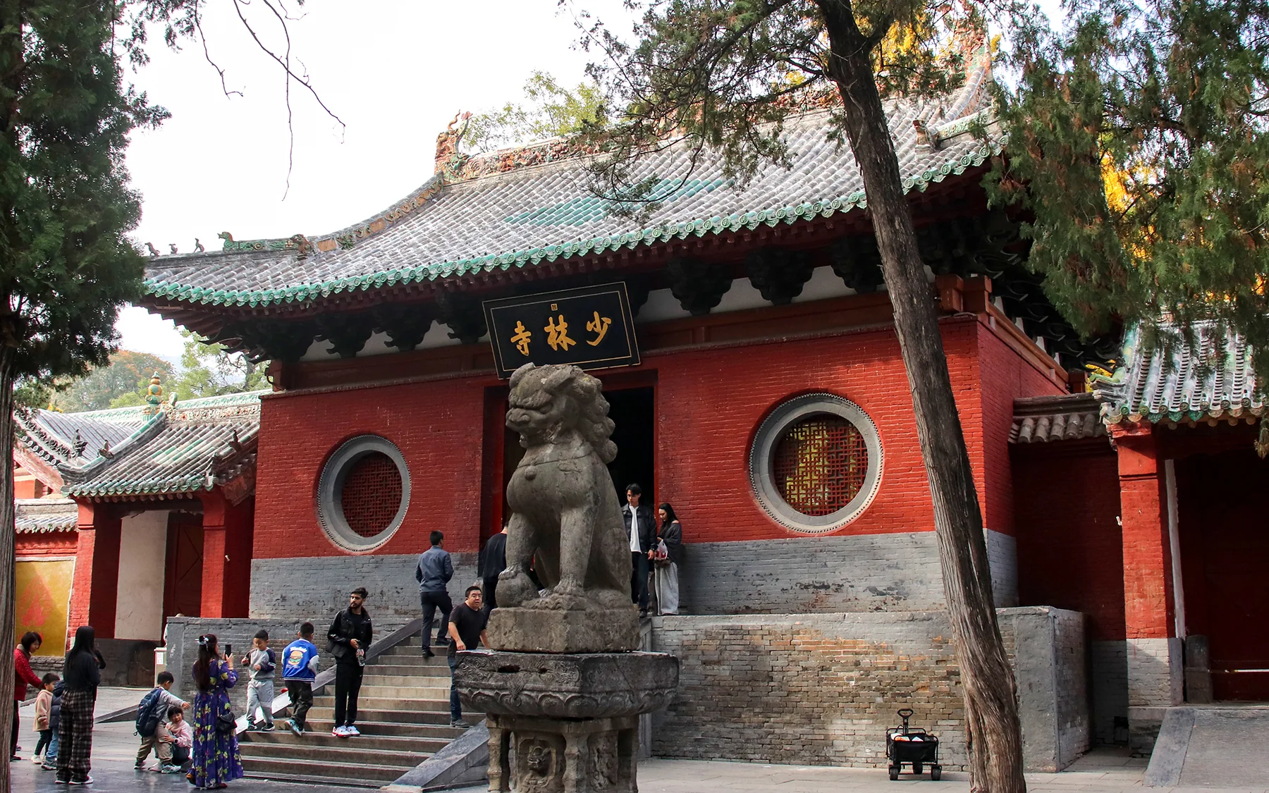 The red mountain gate of Shaolin Temple beneath mature cypress trees, with the gold Shaolin Temple plaque and stone guardian lion clearly visible.
