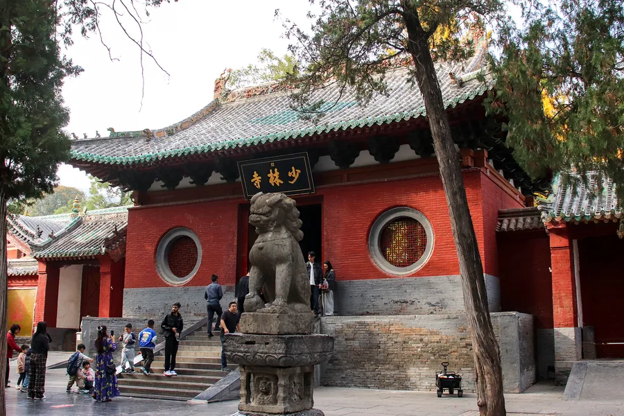 The red mountain gate of Shaolin Temple beneath mature cypress trees, with the gold Shaolin Temple plaque and stone guardian lion clearly visible.