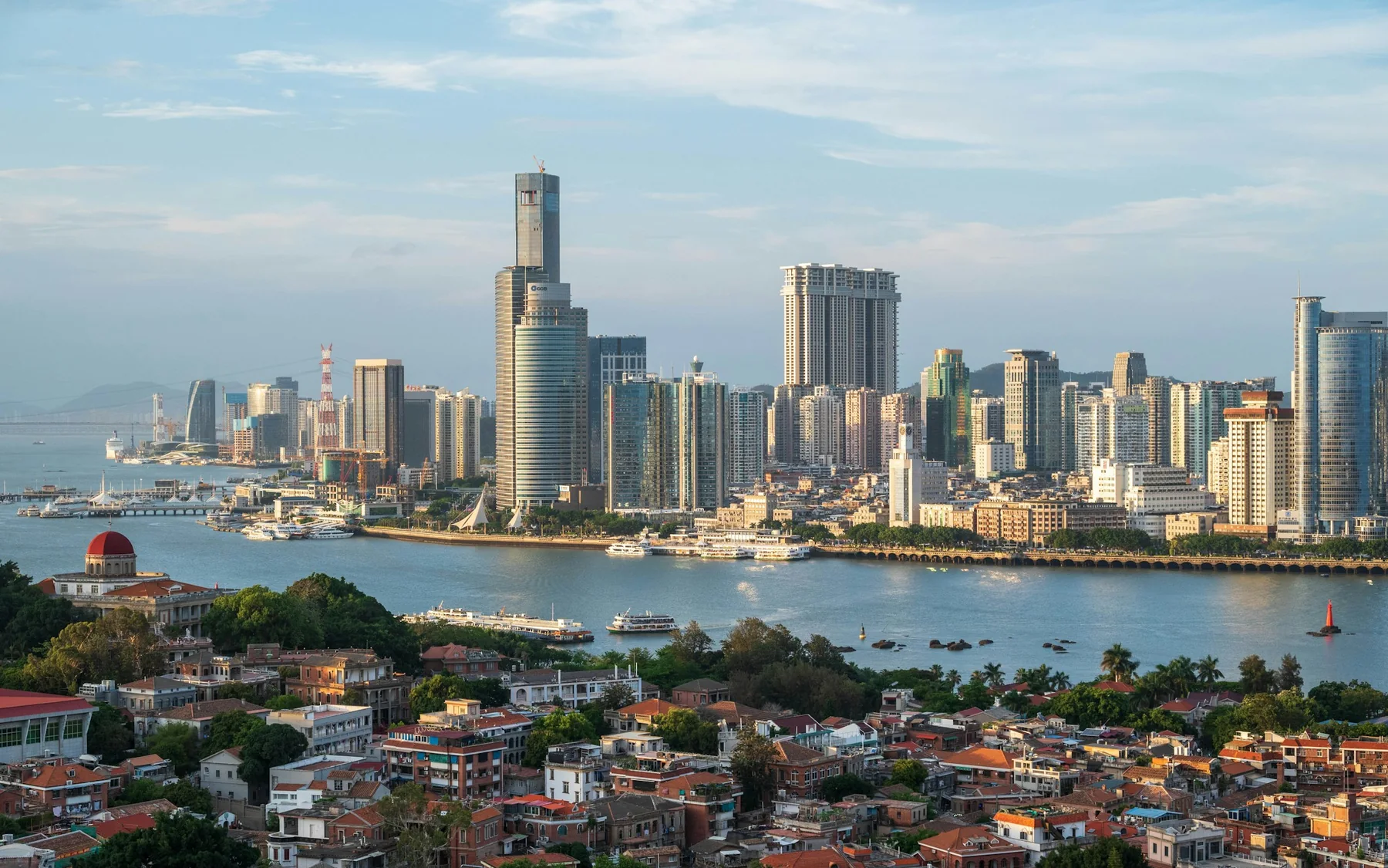Gulangyu's red-roofed historic neighbourhood in the foreground, with ferries crossing toward Xiamen's waterfront skyline in clear daylight