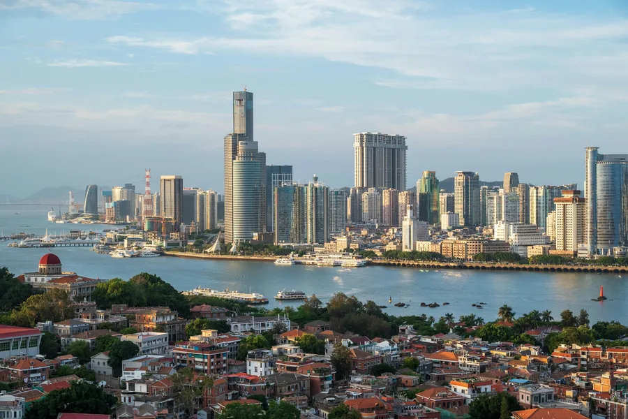 Gulangyu's red-roofed historic neighbourhood in the foreground, with ferries crossing toward Xiamen's waterfront skyline in clear daylight