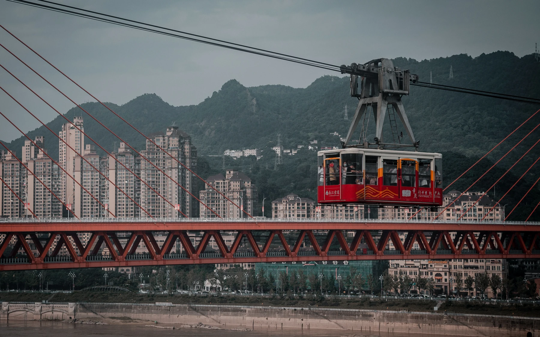 A red Yangtze River Cableway cabin crossing above the riverbank, framed by cables, the red Dongshuimen Bridge and Chongqing's steep urban hillside.