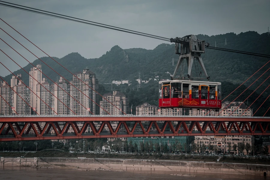 A red Yangtze River Cableway cabin crossing above the riverbank, framed by cables, the red Dongshuimen Bridge and Chongqing's steep urban hillside.
